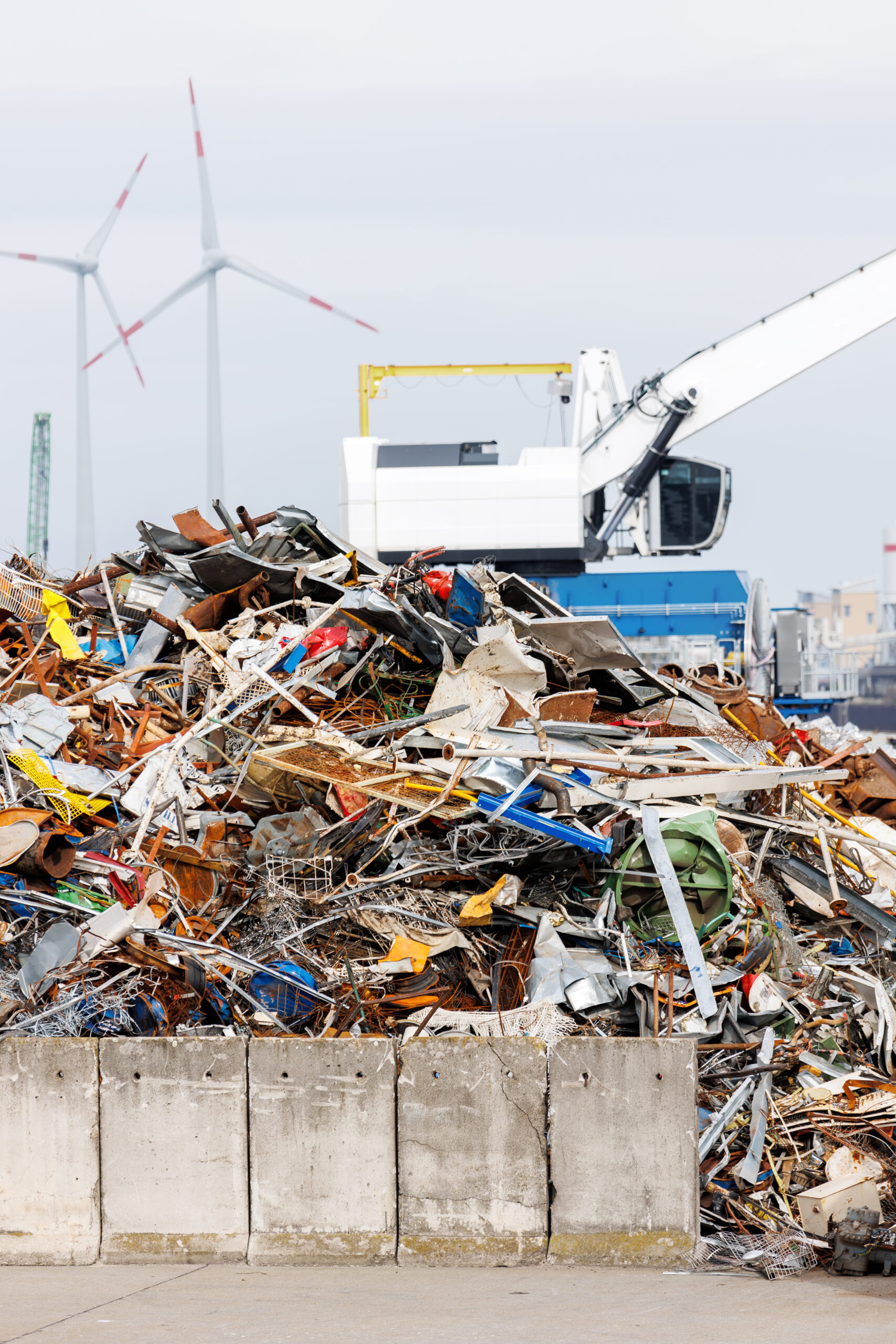 Crane at recycling facility loading large scrap metal pile with wind turbines in background. Industrial area scene waste management and resource reuse concept